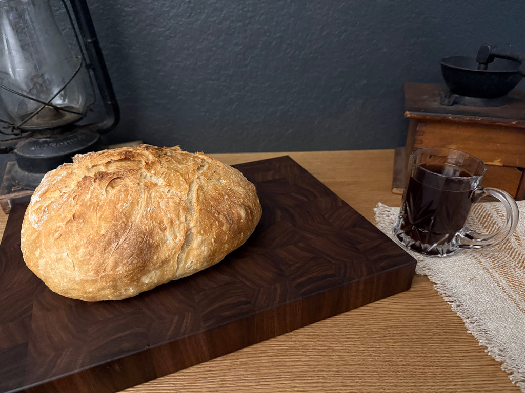 A rustic round loaf of bread sits on a dark wood cutting board on a wooden table, with a glass mug of dark beverage and a lantern in the background.