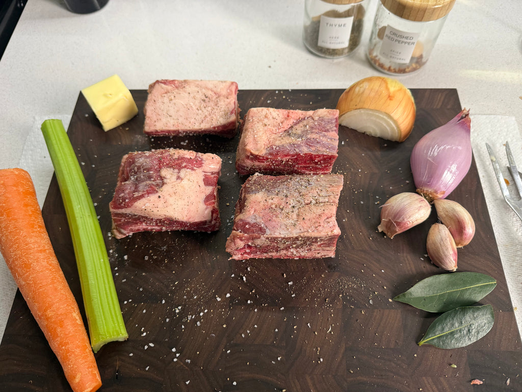 Four raw beef short ribs seasoned with coarse salt and pepper, arranged on a dark wooden cutting board with raw vegetables including a carrot, celery, onion, shallot, garlic, and bay leaves, with spice jars in the background.