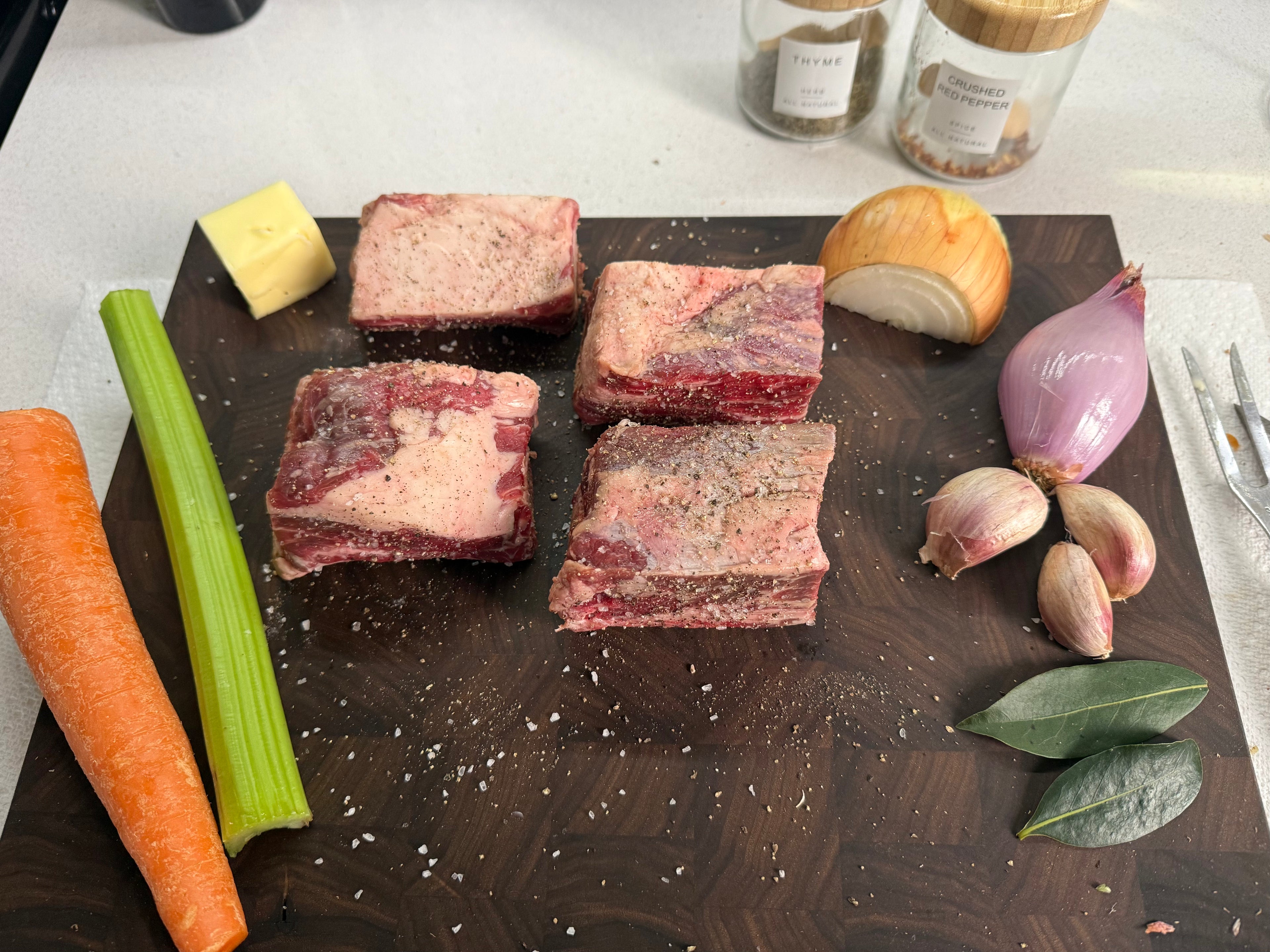 Four raw beef short ribs seasoned with coarse salt and pepper, arranged on a dark wooden cutting board with raw vegetables including a carrot, celery, onion, shallot, garlic, and bay leaves, with spice jars in the background.